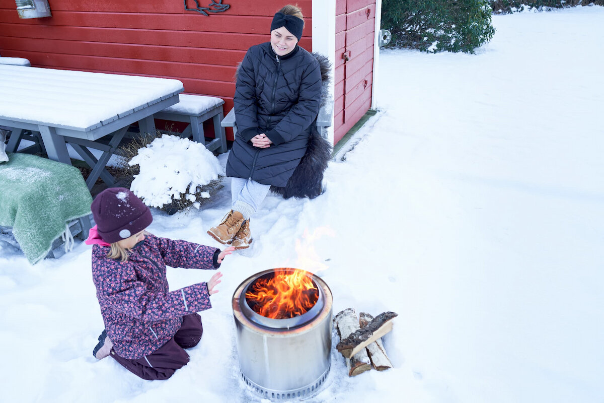 Kvinne og barn sitter rundt en utepeis om vinteren